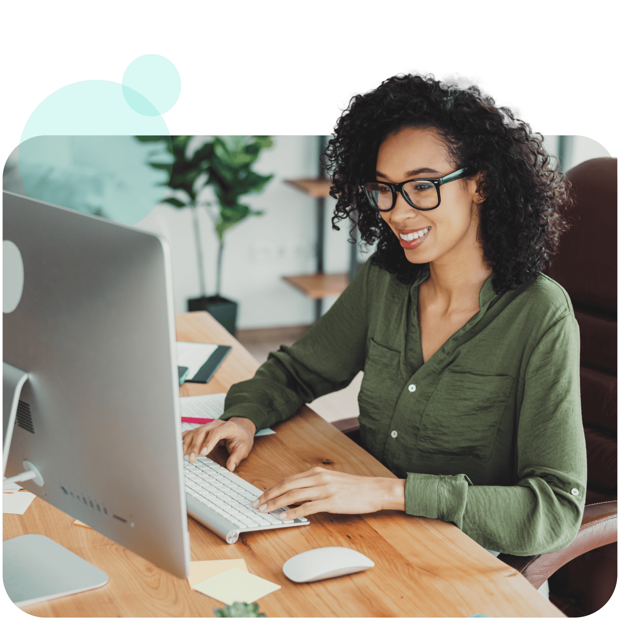A woman with curly hair and glasses is sitting at a wooden desk, smiling while working on a large desktop computer. She is wearing a green shirt and typing on a keyboard, with office supplies and plants visible in the background.
